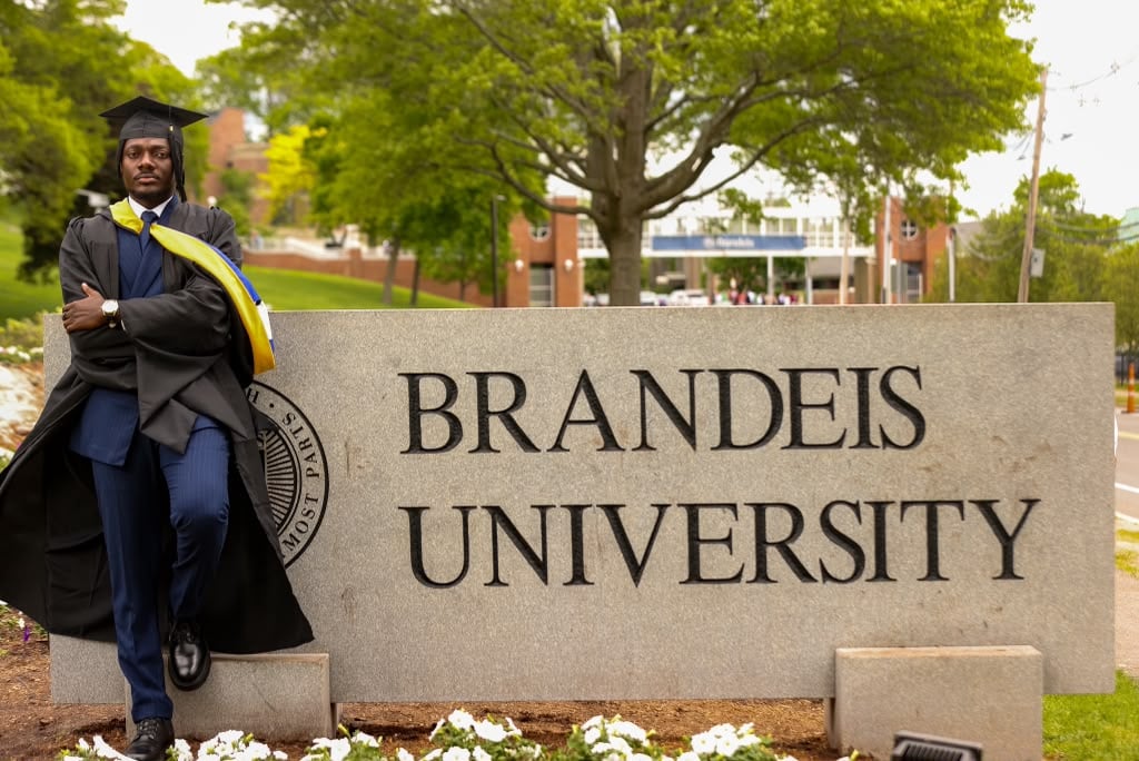 Terrick in cap and gown leaning against the Brandeis University sign on graduation day